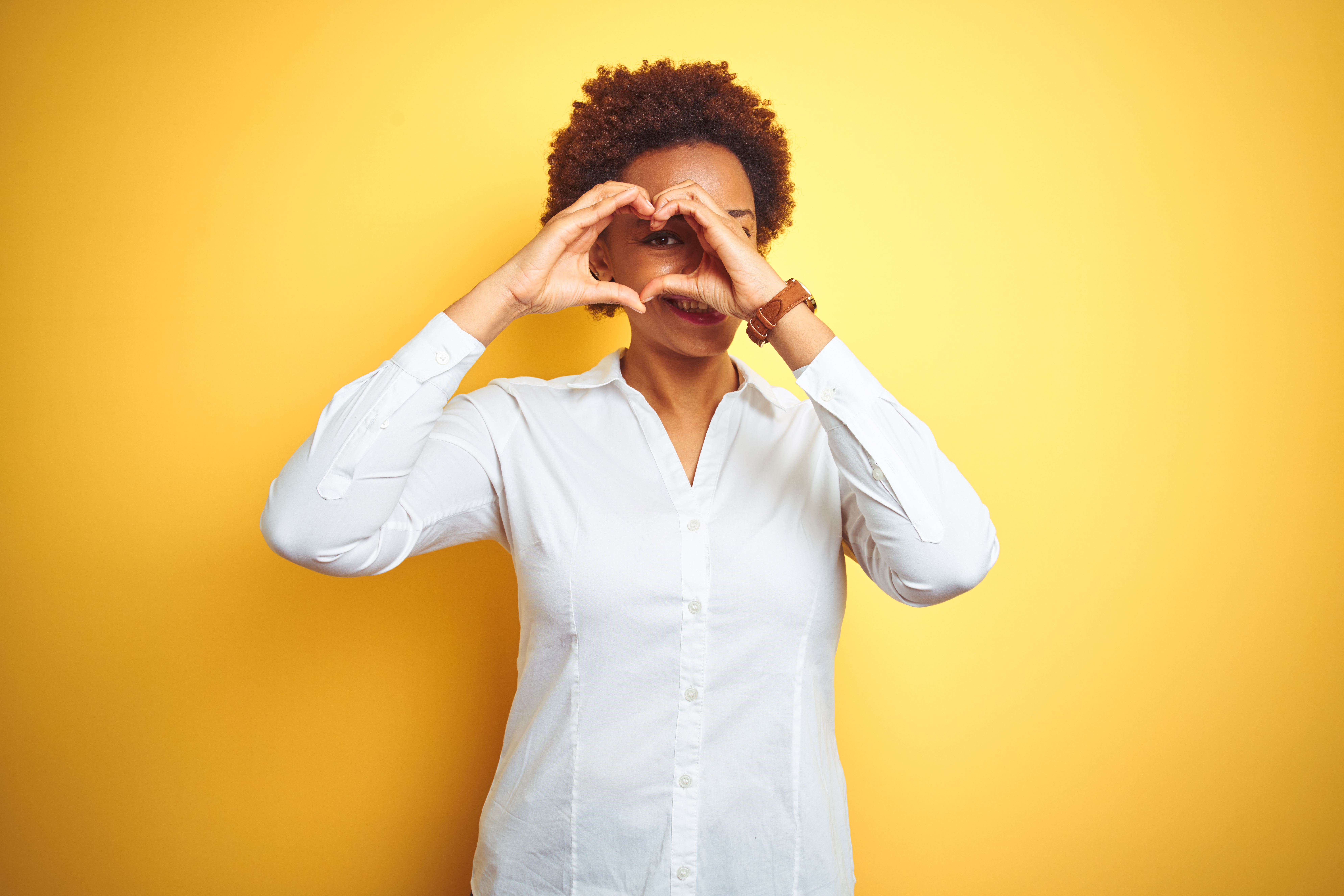 African american business woman over isolated yellow background Doing heart shape with hand and fingers smiling looking through sign African American woman making heart symbol with her hands in front of her eyes